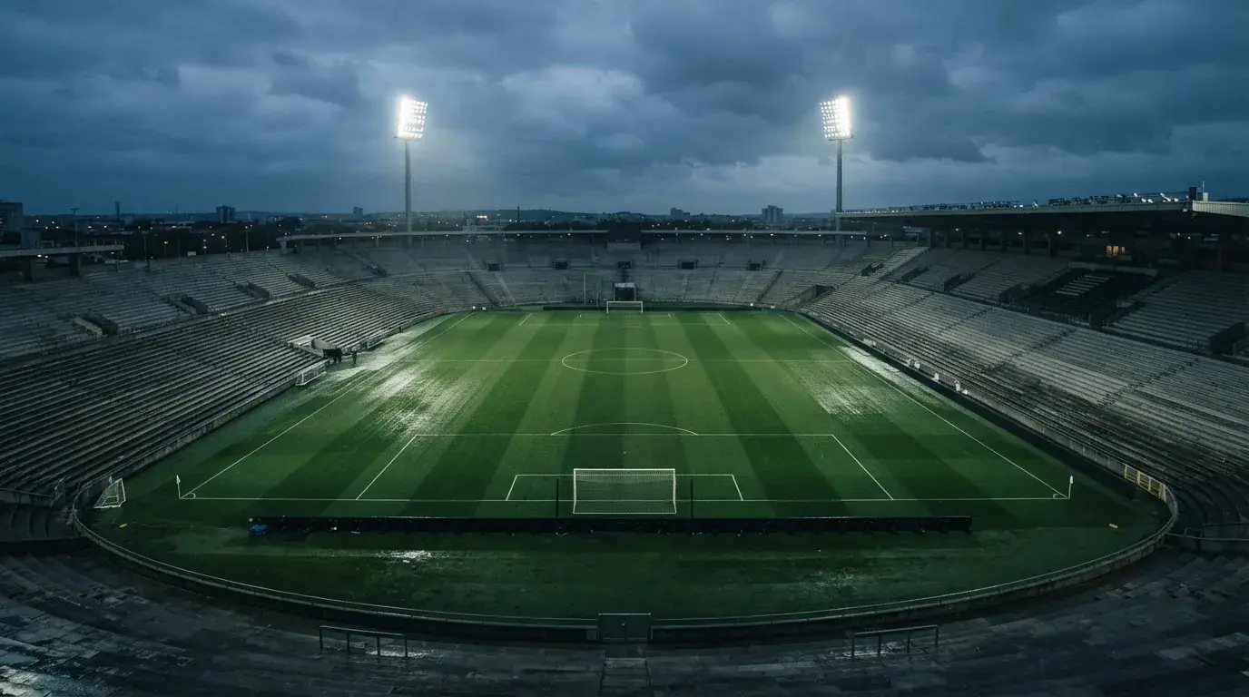 Ein Fußballplatz mit Flutlichtmasten in der Abenddämmerung, aus der Ferne gesehen, leere Tribünen und nasser Rasen unter bewölktem Himmel.