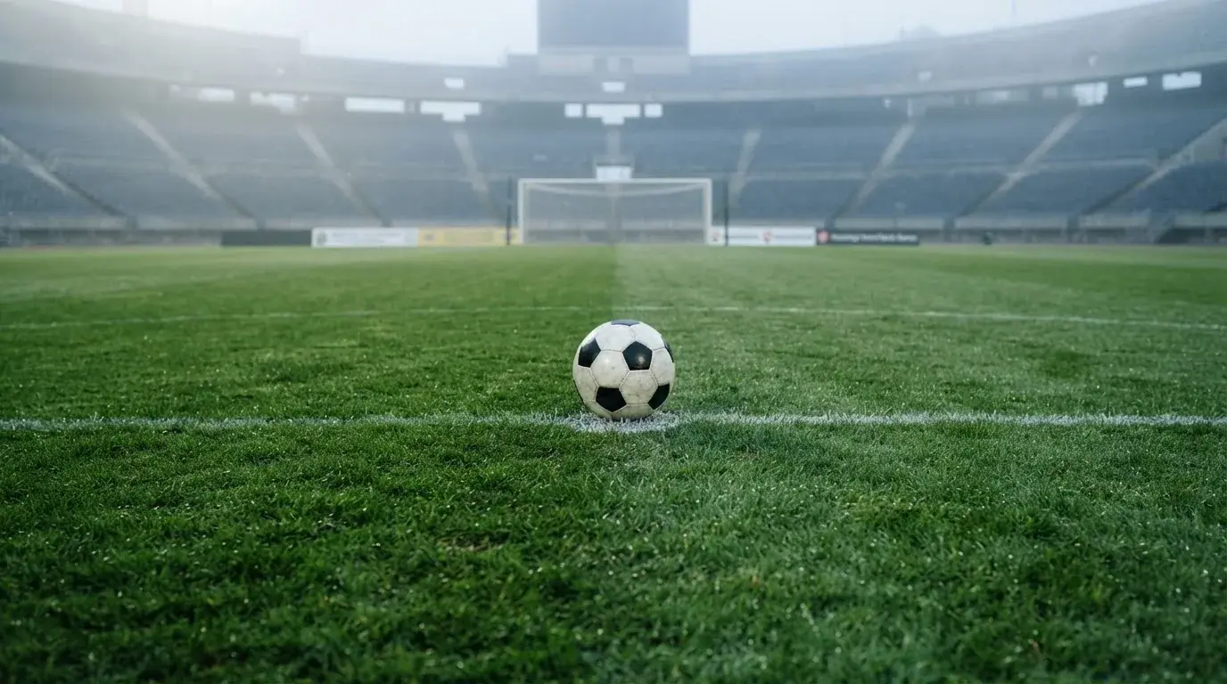 Ein Fußball liegt auf dem Elfmeterpunkt eines leeren Rasenplatzes, im Hintergrund eine Tribüne im Dunst.
