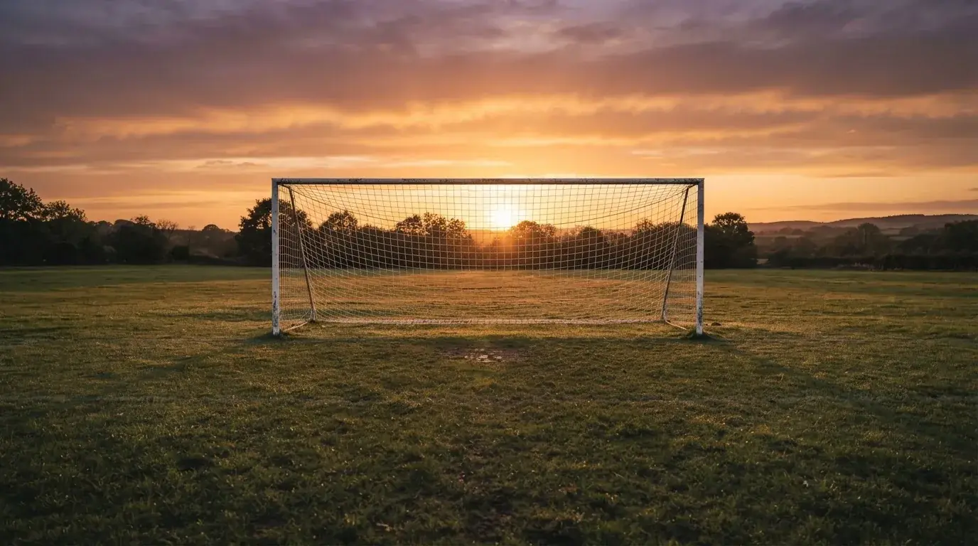 Ein Fußballtor mit Netz bei Sonnenuntergang auf einem leeren Platz, die Schatten der Torpfosten liegen lang auf dem Gras.