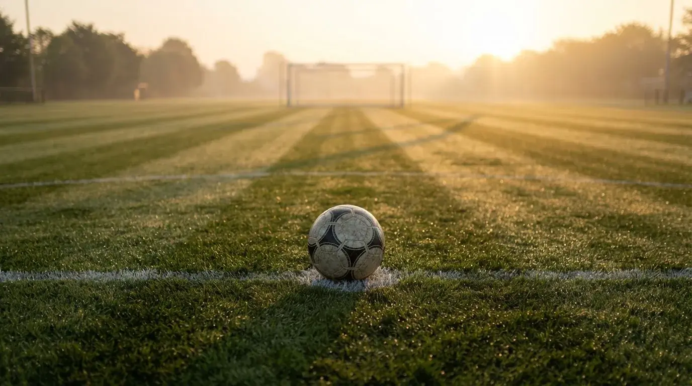 Ein Fußball liegt auf dem Anstoßpunkt eines Rasens bei Sonnenaufgang, Tau glitzert auf den Halmen.