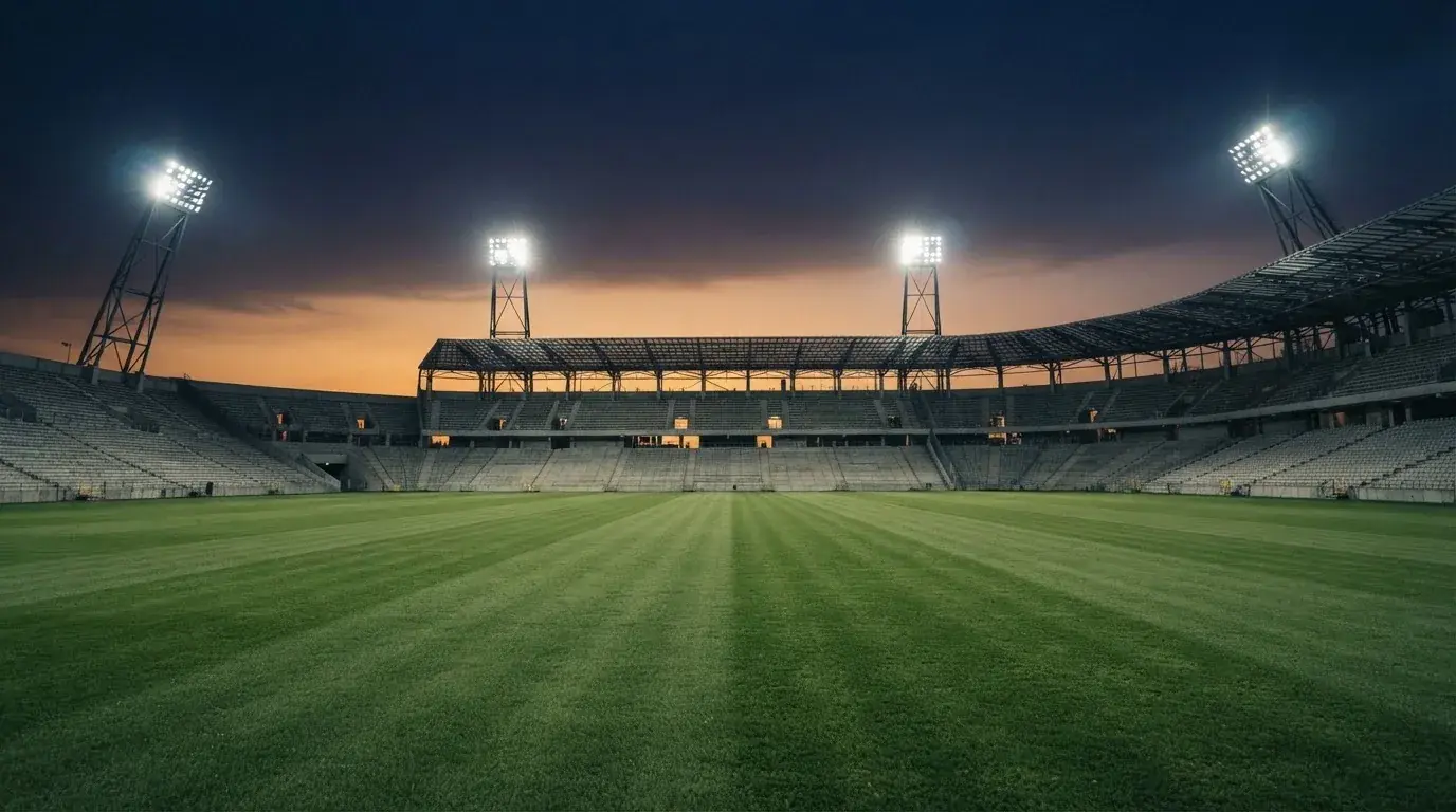 Eine leere Stadiontribüne unter Flutlicht aus der Ferne betrachtet, die Rasenfläche leuchtet grün in der Dämmerung.