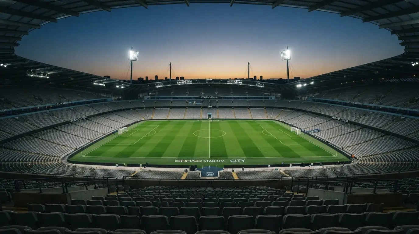 Eine leere Stadiontribüne unter Flutlicht aus der Ferne betrachtet, die Rasenfläche leuchtet grün in der Dämmerung.
