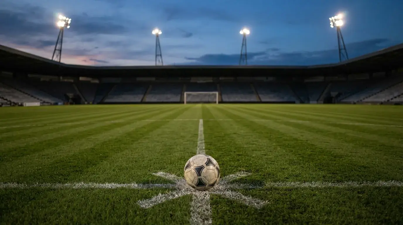 Ein Fußball liegt auf dem Rasen eines leeren Platzes, im Hintergrund ein Stadion unter Flutlicht in der Abenddämmerung.