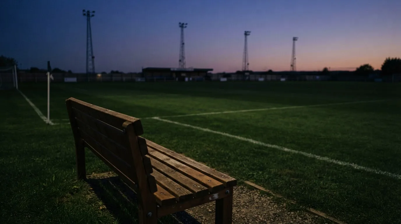 Eine leere Parkbank am Rand eines Fußballfelds in der Abenddämmerung, im Hintergrund ragen dunkle Flutlichtmasten in den Himmel.