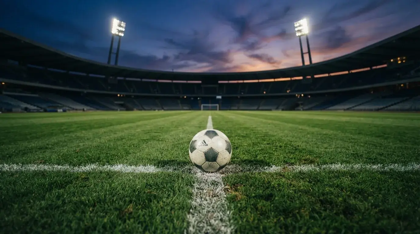 Ein Fußball liegt auf dem Anstoßpunkt eines leeren Rasenplatzes, im Hintergrund steht ein Stadion unter Flutlicht in der Dämmerung.